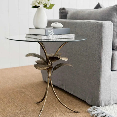 Amari Gold Leaf Side Table styled beside a sofa, featuring a glass top and elegant botanical-inspired base in a warm gold tone.