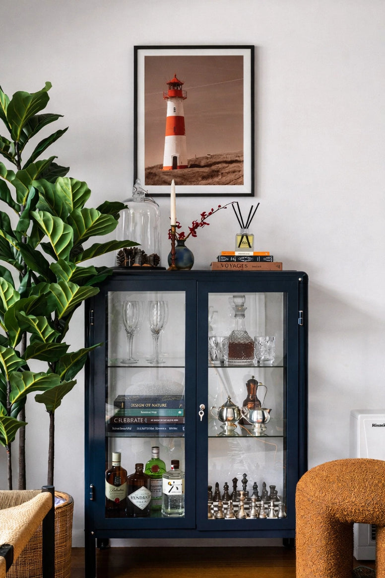 Decorative cabinet with glass doors displaying bottles and glasses, plant to the left, and framed picture on the wall, modern look at Essencia Interiors
